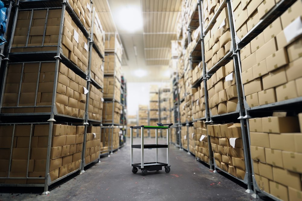Model C2 Autonomous Mobile Robot (AMR) for material transport and item movement stands in the center of a warehouse filled with rows of shelves and boxes