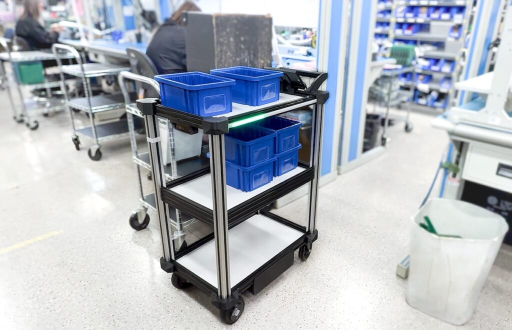 The Model C2 autonomous robotic delivery cart is pictures centered amidst a tight aisle of an electronics manufacturing facility. it is loaded with blue organization bins. the background features racks of reels for electrical components and circuit board assembly.