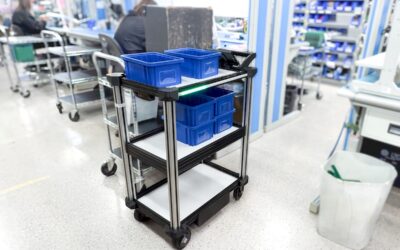 The Model C2 autonomous robotic delivery cart is pictures centered amidst a tight aisle of an electronics manufacturing facility. it is loaded with blue organization bins. the background features racks of reels for electrical components and circuit board assembly.